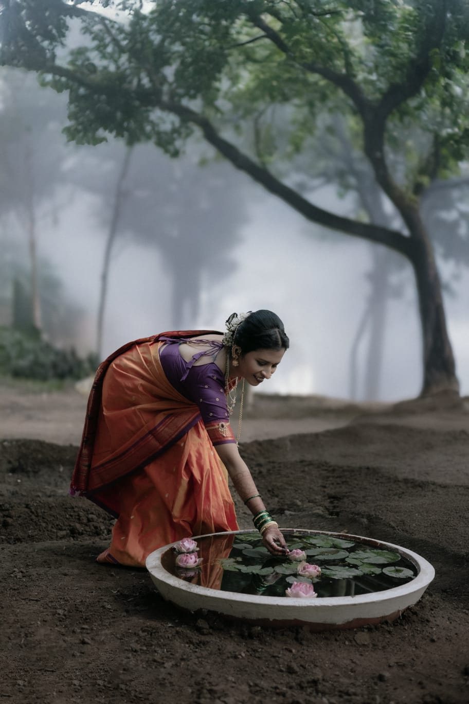 Woman in an orange Paithani saree placing lotus flowers in misty woods